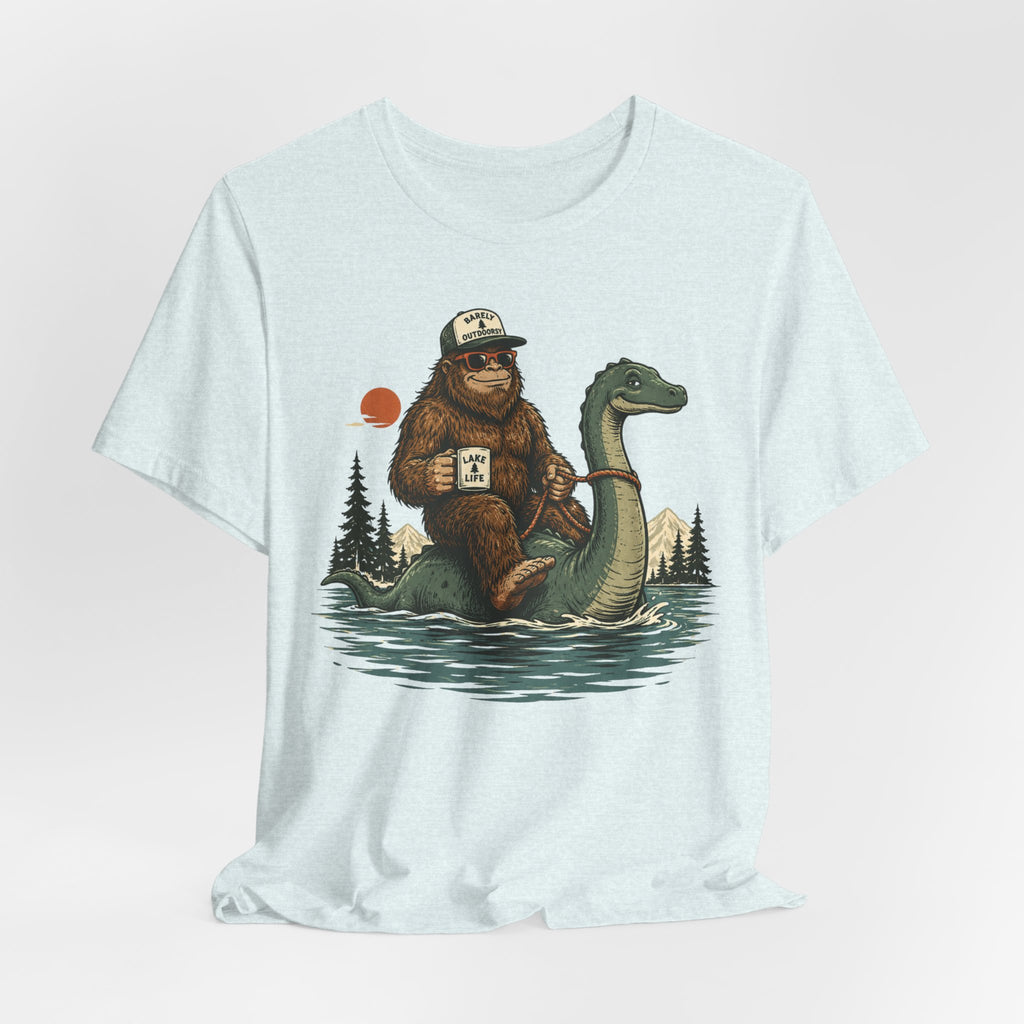 Heather ice blue unisex t-shirt with Bigfoot in Barely Outdoorsy trucker hat and sunglasses riding Loch Ness Monster on lake holding Lake Life mug graphic, Bella Canvas 3001
