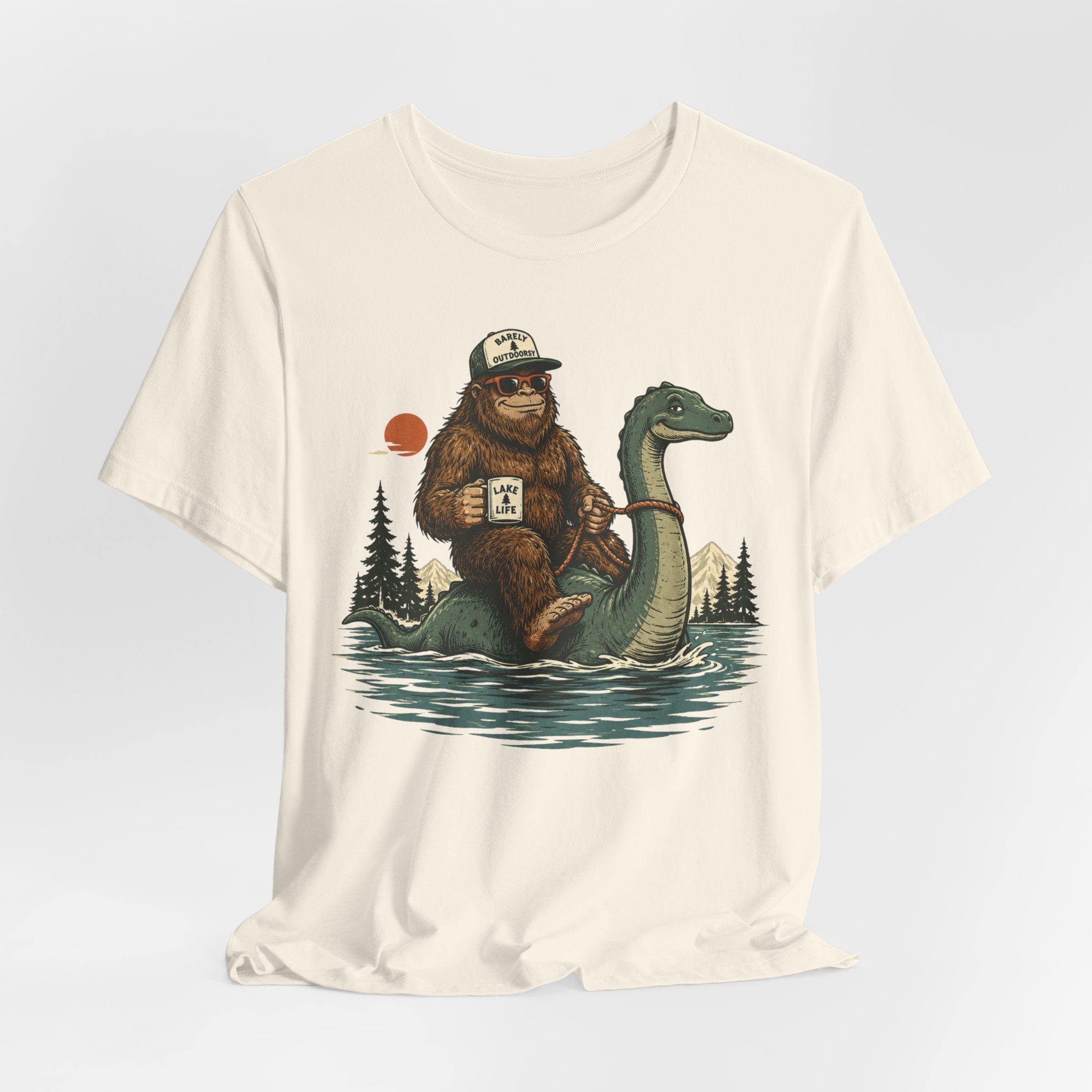 Person wearing heather ice blue Bigfoot riding Loch Ness Monster lake life graphic tee, funny cryptid lake shirt